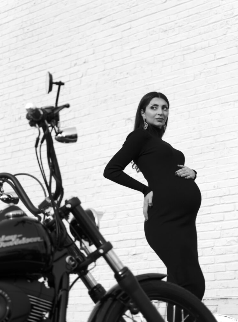Mother to be 2 Portrait of pregnant woman (mother-to-be) standing next to a motorcycle and white brick wall wearing a black dress in NJ.